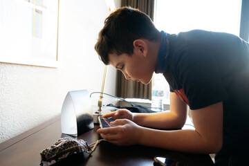 Boy leaned over a desk playing a game on his phone. 