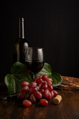 View of bottle and glass of red wine with red grapes, leaves and cork stopper, on wooden table,...
