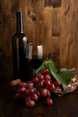 View of bottle and glass of red wine with red grapes, leaves and cork stopper, on wooden table and...