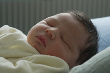 A newborn baby in a soft yellow onesie sleeps peacefully on a blue pillow, with tiny hands resting...