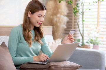 Young asian woman sitting on sofa using laptop computer shopping online with credit card buying to internet, female payment with business on couch, purchase and payment, business and lifestyles.