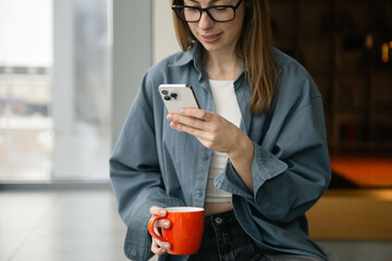 A woman is using a smartphone and holding a cup while sitting by the window.