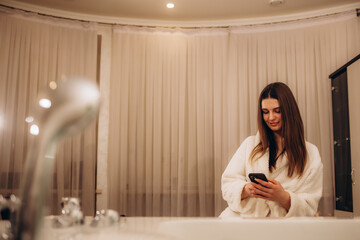 Smiling young indian woman in white bathrobe using smartphone after shower, sitting on bathtub in...