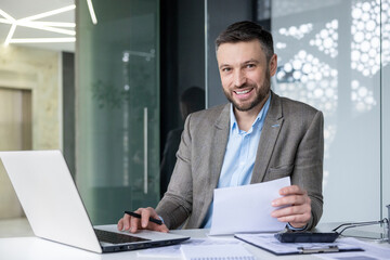 Smiling businessman at his desk working on laptop and checking documents in a contemporary office setting.