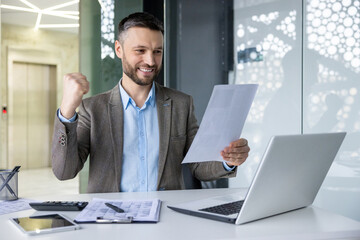 Fototapeta premium Cheerful businessman celebrating achievement in modern office environment, looking at documents while working on laptop