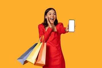 A joyous young woman dressed in a vibrant red dress stands against an orange background, holding colorful shopping bags in one hand and presenting a blank-screen smartphone mockup