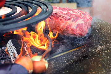 A large pieces of raw fresh steak are grilling on flaming grill.