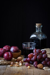 View of rustic bottle and glass with red wine, grapes, plums and nuts on wooden table, black...