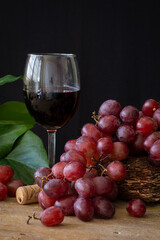 Close-up of glass with red wine, grapes, cork and green leaves on wooden table, black background, vertical, with copy space