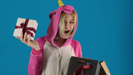 Female teenager in unicorn onesie takes gift box out of shopping bag on blue background. Girl expresses joy with surprise