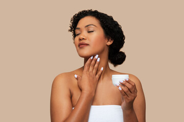 A young woman with curly hair, wrapped in a white towel, gently applies moisturizing cream to her neck, moment of self-care and relaxation. The neutral beige background
