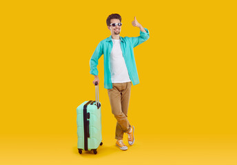 Passenger who likes to travel. Happy joyful man who bought cheap flight tickets shows thumbs up while posing with travel suitcase. Guy in casual clothes and glasses on yellow background. Full length.
