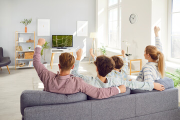 Back view of joyful young family fans sitting on sofa in living room and watching football match on...