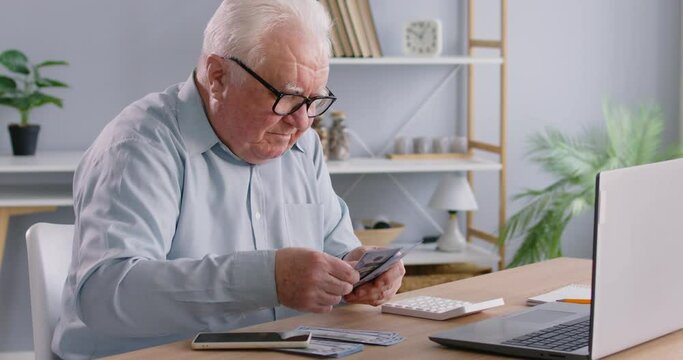 Portrait of grey-haired senior old man counting money cash sitting at the desk with laptop at home. Elderly retired person calculating income and profit. Pension calculation concept. 4k video.