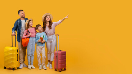 A happy family consisting of a father, mother, son, and daughter stands with their colorful suitcases, prepared to embark on a vacation, pointing at copy space