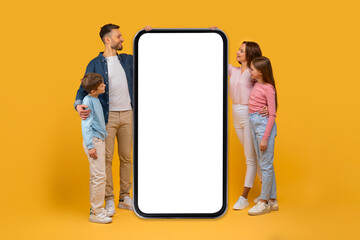 A family consisting of a man, woman, boy, and girl are standing together beside an oversized smartphone with a blank white screen, suitable for mockup purposes.