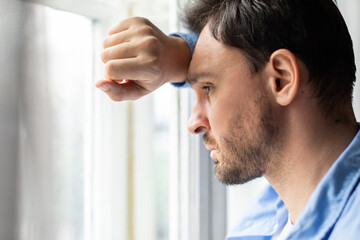 A man rests his forehead on his hand while looking thoughtfully out of a bright window, his...