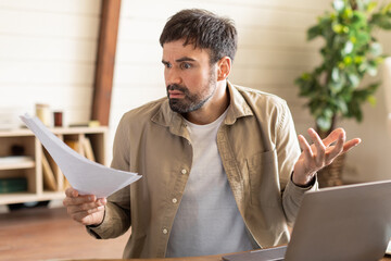 A man with a beard exhibits a puzzled expression as he examines papers in home office, laptop is...