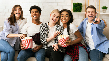 A diverse group of five young friends is seated comfortably on a sofa, sharing laughter and food....
