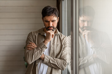A contemplative man leaning on a large window pane, with his reflection visible. He appears deep in...