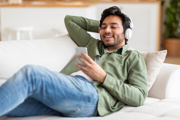 Arab man is seated on a couch, attentively listening to music playing from his phone, using...