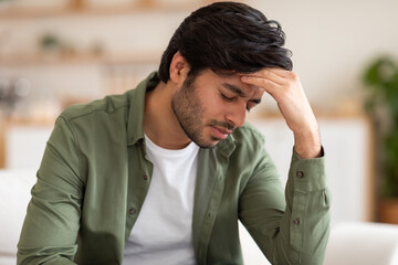 Arab man is seated on a couch, leaning back with his hand on his head. He appears deep in thought or contemplation, with a pensive expression on his face