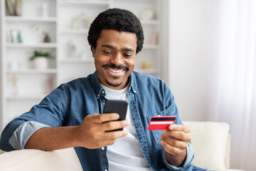 African American man is seated on a couch, holding a smart phone in one hand and a credit card in...