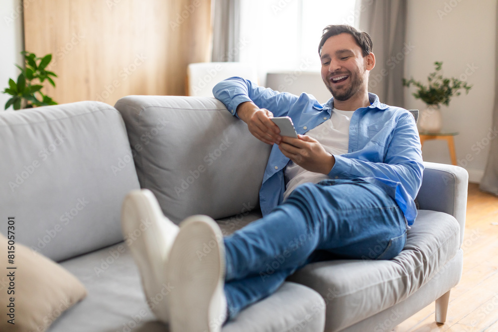 Poster a man is seated comfortably on a couch, engrossed in his phone. he is scrolling and typing, complete
