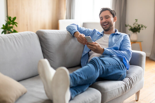 A man is seated comfortably on a couch, engrossed in his phone. He is scrolling and typing, completely absorbed in the digital device in his hands.