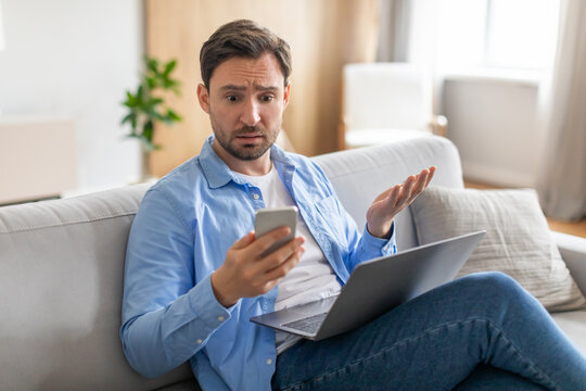 A man sits on a couch with a laptop on his thighs, holding a smartphone in one hand while displaying a puzzled expression, appears to be encountering unexpected news or a perplexing situation