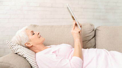 Senior woman is lying on top of a couch indoors, holding a tablet in her hands. She appears relaxed and focused while browsing or interacting with the device, reading blog