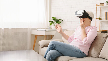 Senior woman is seated on a couch, fully immersed in a virtual reality experience as she wears a...