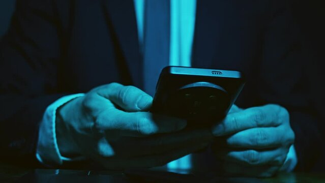 Hands of man in business suit using mobile phone in the dark sitting at table. Dark blue composition scheme