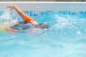 Child swimming freestyle, creating splashes in a sunny outdoor pool.