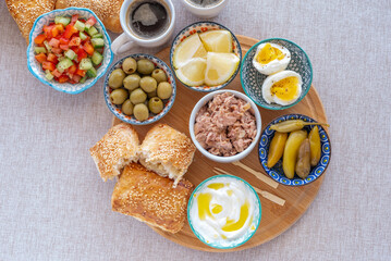 Israeli breakfast including traditional pastry Bourekas, green olives, vegetable salad, yoghurt, pickled peppers, boiled eggs, tuna served on wooden tray over cotton fabric background.
