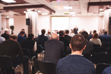 A large group of people are sitting in a room, listening to a speaker. Scene is serious and focused, as everyone is attentive to the speaker