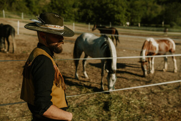 Rancher in hat standing near fenced pasture with horses graze on it.