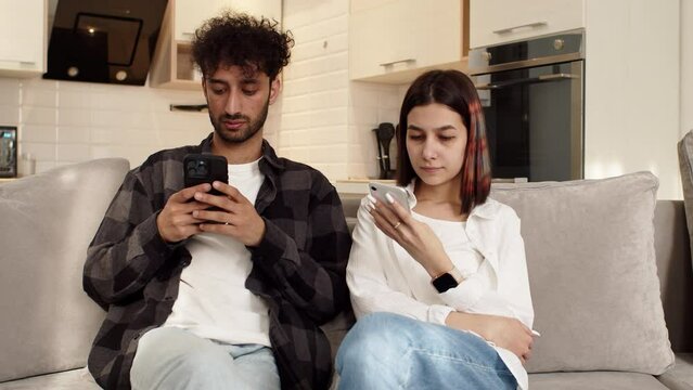 Young couple sitting on the couch at home with their smartphones and don't pay attention to each other. Concept of difficulties in a couple relationship