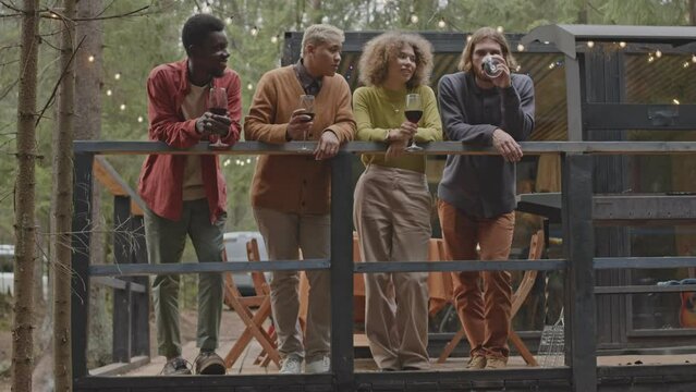Company of young diverse friends drinking wine, looking at nature and chatting while standing on terrace of cabin in forest decorated with string lights