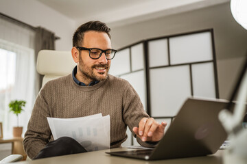 Adult man sit at office look on laptop and hold paper at office