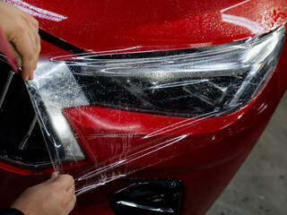 The master applies vinyl film to the headlight of a red car. 