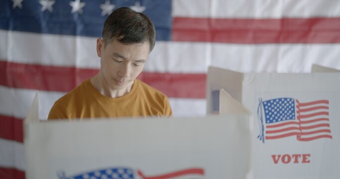 Frontal close up on Asian man casting votes in American election booth. Empty booth and US flag soft focus in background