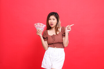 expression of a sad indonesia woman holding rupiah money while pointing to the left wearing a brown...