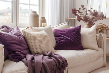 Beige sofa with violet pillows and chunky knit plaid near window. Scandinavian hygge interior design of modern living room home.