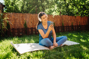 One young caucasian woman is sitting on yoga mat with wireless headphones and mobile phone preparing for training	