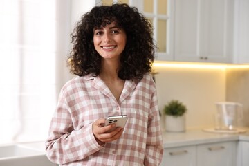 Beautiful young woman in stylish pyjama with smartphone in kitchen