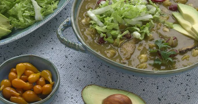 Vegan Pozole Verde dish topped with shredded napa cabbage, red radish and avocado. More toppings on the table. Table spin.