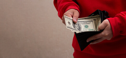 Man in red hoodie pulls out 100 USD. Dollar bill from black leather wallet. Horizontal banner with...
