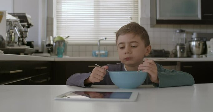A young boy is seated at a modern kitchen table, focused on a digital tablet with a bowl of cereal in front of him.