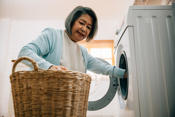 An old woman, a grandmother, loads laundry into the washing machine, smiling as she covers her...